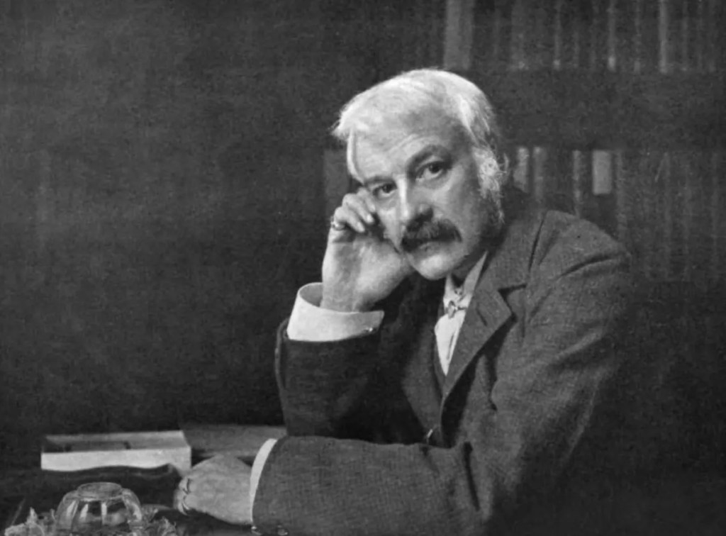 A black and white portrait of a thoughtful man with a mustache, dressed in a suit, resting his chin on his hand, sitting at a desk with books in the background.
