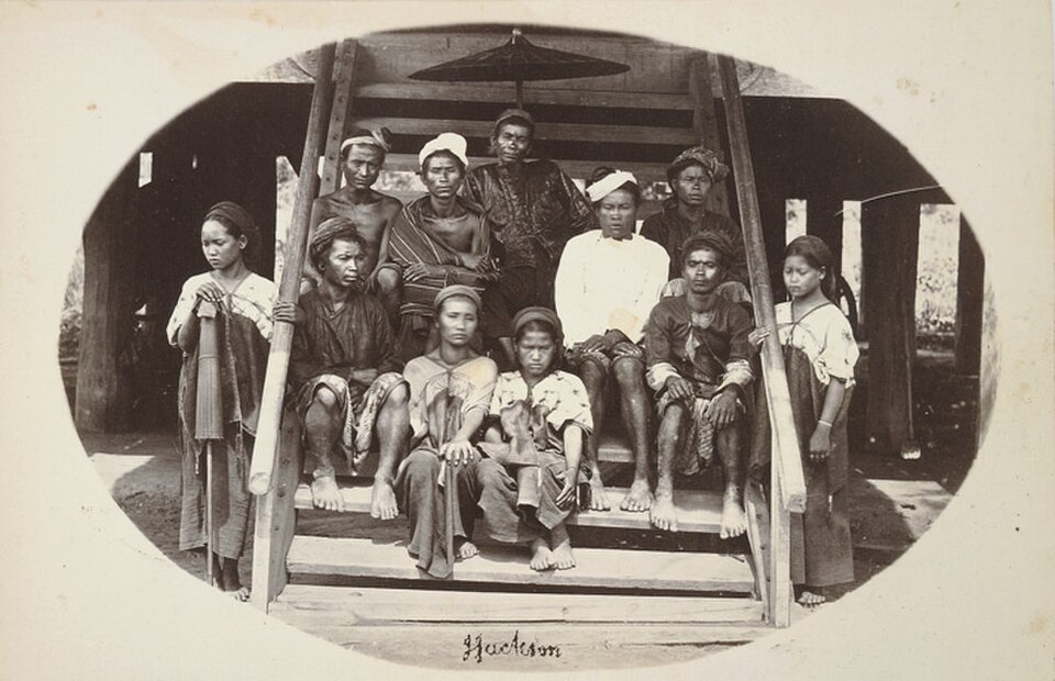 A historical black and white photograph featuring a group of eleven individuals sitting on steps, dressed in traditional attire, with some wearing headscarves and decorative accessories.